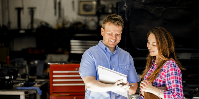 Happy customer!  A smiling Latin descent woman discusses automobile repair invoice with an auto mechanic in a repair shop.  She is discussing the vehicle's repairs with the mechanic, who is explaining her service.  Toolbox, workshop background. Unidentifiable, SUV-style vehicle.